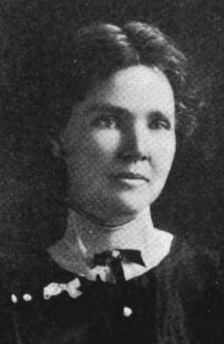 A white woman with dark hair, center parted and dressed back to the nape, wearing a high-collared blouse with a dark overdress