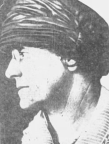 An older woman in profile, wearing a turban-style hat and glasses