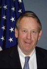 Smiling man with thinning hair wearing a suit and a blue tie with the US flag behind him