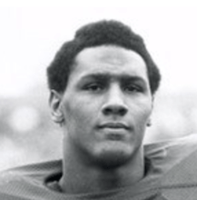 tight headshot of a dark skinned man with a slightly long afro lookin into the camera. His football shoulder pads are slightly visible.