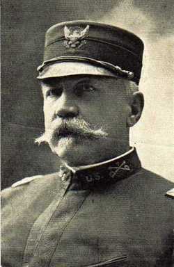 Black and white picture of Louis Carpenter, a white male with a large mustache wearing his US Army uniform. His Army uniform has a high collar with an emblem of crossed swords and the letters U.S. next to it. He is wearing a round hat with an emblem of an eagle clutching something.
