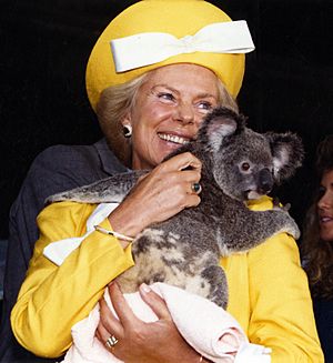 The Duchess, smiling and in bright yellow clothes, holds a koala