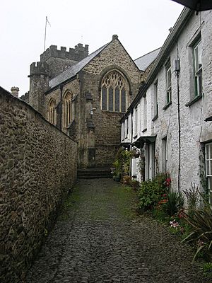 Stone building with arched windows and square tower seen at the end of a narrow lane with white painted houses on the right and a wall on the left
