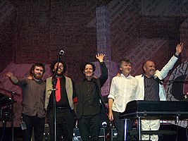 Five men are standing close together on a stage and smiling. Behind the five men is more band equipment and the background contains considerable English text.