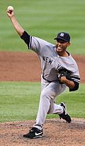 Mariano Rivera in a gray baseball uniform and navy blue cap stands on a dirt mound. He is striding forward to the left as he holds a baseball in his forward-extended right arm. His face is contorted in concentration.
