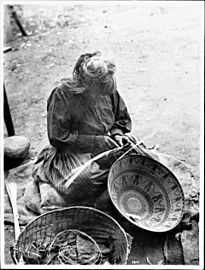 Woman weaving a wide, shallow, patterned basket while sitting on the ground