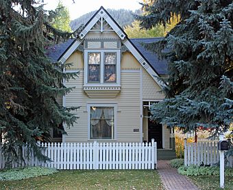 An ornate pale yellow wooden house with a pointed roofline between two tall evergreen trees. There is a white picket fence in front and a distant ridgeline in the rear.