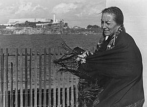 A photograph of a woman standing behind a fence, opposite of Alcatraz island. She is wearing a dark tassled shawl, which is blowing in the wind and her long hair is braided with beads, leather straps, and animal fur.