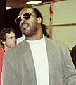 Stevie Wonder, a middle-aged African American man with his hair tied in a ponytail, dark sunglasses and stubble on his face. He wears a plain white tee-shirt and a grey jacket.