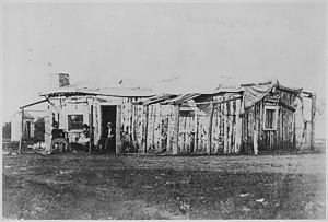 "Officers quarters at Ft. Rawlins, Wyoming, May 7, 1877." Two men and a woman holding a baby sit in front of the crude structure - NARA - 531107