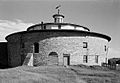 This black and white photo shows a round barn made of fieldstone. It is topped by a white wood frame multi-sided section that is smaller, and then a small turret.