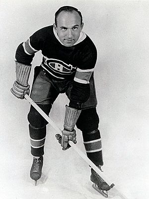 An ice hockey player leans forward with his stick. He has a round face with one eyebrow raised and a bare head with a receding hairline. He wears skates, gauntlets, and a sweater with a stylized "C" around a smaller "H".