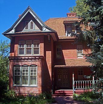 An ornate brick house with a gabled section projecting at the left and a hipped roof on the main section behind it. The doors, windows and porch have detailed wooden trim painted red, yellow and green.