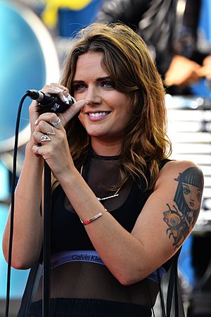 A woman with long brown hair wearing a black top and holding a microphone stand with both hands