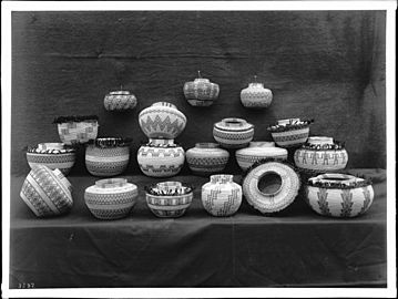 A collection of 19 unidentified Indian baskets on display, photographed ca.1900. They are displayed on two tiers covered with a cloth and displayed hanging from a blanket backdrop. They mostly have the shape of a round bowl with a narrow neck and mouth. They have differing woven patterns including stripes, zig-zags, diamonds, steps, human figures and more. Many of them have tassels around the widest part of their bulge.