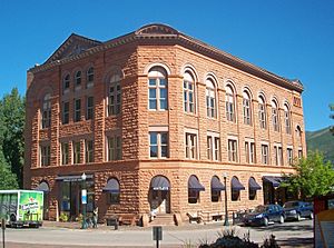 A three-story orange stone building on a slightly curved street corner, lit by sun from the right. The windows at street level have awnings; those at the top are in rounded arches. The face of the building on the left has a small pointed top at the roof. At street level an awning on that face has "Wheeler Opera House" on it; the word "Bank" is carved into the stone on the corner face above an awning with "Bentley's" written on it.