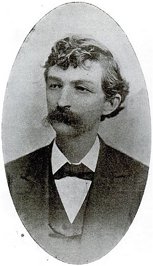 A man with curly, dark hair and a thick, dark mustache wearing a black jacket and tie and white shirt