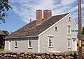 A gray saltbox with white trim is seen at an angle from the rear. A low stone wall stands in the foreground.