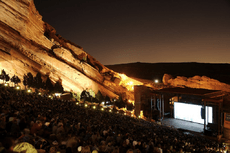 A shot of an outdoor amphitheatre taken at dusk, looking down towards a brightly lit stage. Large red cliffs are visible in the background, sloping down to the right. Several hundred people are visible between the camera and the stage.