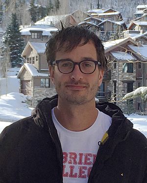David Farrier wearing dark jacket with white t-shirt with lettering underneath, looking directly at camera, with snowy urban landscape as a backdrop