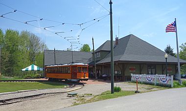 The Visitor Center is a large green building with a high peaked roof styled after a train station, with a waiting room inside and a boarding platform outside. An orange electrically-powered streetcar from the Boston Elevated Railway is on the loop track in front of the Visitor Center.