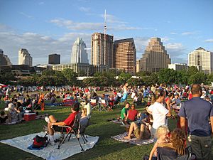 Slightly elevated image taken during the evening of a crowd of people relaxing in a park, with a skyline visible in the background.