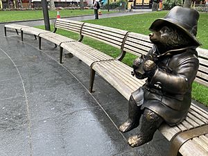 A statue of paddington bear, sitting alone on a bench while looking happy eating his marmalade sandwich. The bear is a cub, and is wearing a raincoat and a big hat.