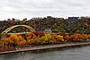 Red, orange, and yellow leaves on trees in an urban park on the shore of a river, a yellow bridge rises in the background