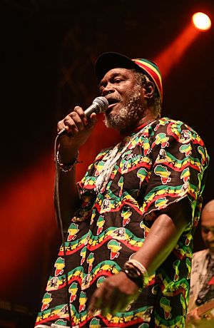 Horace Andy wearing a Rastafarian-patterned shirt and hat, standing onstage, singing into a microphone