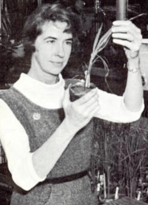 A white woman wearing a tweedy jumper-dress and a white turtleneck; she is holding a small potted plant in one hand, and holding one of its leaves with the other hand