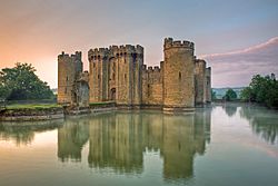 Photo of Bodiam Castle at sunset with towers and battlements reflected in a wide moat. A path leads away from the entrance of the castle across two islands