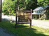 A brown wooden sign with yellow letters reading "Sand Bridge State Park Picnic Area Pennsylvania Department of Conservation and Natural Resources" in front of a highway and pale green two story frame house