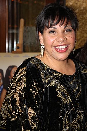 A dark-haired, smiling woman with long, dangling earrings looks right toward the camera