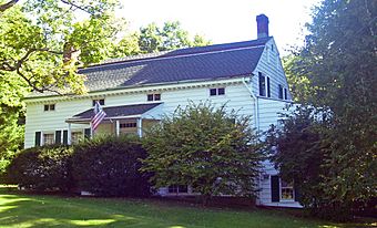 A white house with gray roof and large shrubs in front flying an American flag from the porch