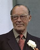 Prince Bernhard is wearing a dark jacket, with a carnation on it, a red tie, grey/brown shirt. He has a slight smile and is wearing spectacles.