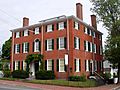 This is a three story red brick house with white trim and black shutters. The third floor has windows that are not as high as those on the first two floors. Three sizable chimneys are visible.