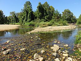 In the foreground is a rocky shallow stream that forks into two a short ways off from the photographer. There is an island in the right fork so that it joins the left fork in two places. Beyond the banks, a grove of deciduous trees rise into blue skies.