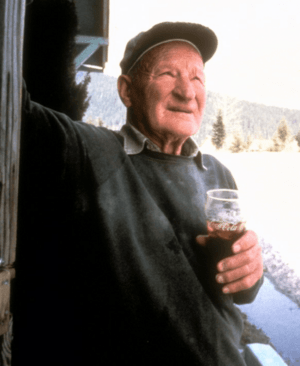 Color picture; An elderly man holding a Coca-Cola glass and wearing a hat leans on the wall of a wooden lodge