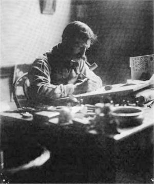 Black-and-white photograph of a man with a large mustache and a pipe in his mouth, at work, seated behind a cluttered desk on which a drawing board is propped at a slight angle.