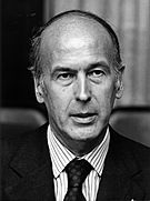 A candid head shot of Valéry Giscard d'Estaing who is looking neutral, wearing a dark jacket, dark tie and a white formal shirt with darker vertical stripes.