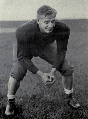 Posed black and white photograph of Petoskey wearing a dark-colored football uniform without a helmet and standing on a grass field in a two-point football stance
