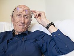 close-up of Stewart Brand wearing a dark blue shirt, holding his glasses up slightly above his eyes, smiling and looking left of camera