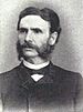 Head and shoulders of a white man with bushy sideburns connecting to a mustache, wearing a suit coat and bow tie.