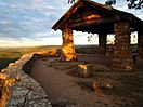 A small, square fieldstone shelter with a roof at the end of a hiking trail. Looking over the edge of the fieldstone retaining wall is the dark green treetops of the Ozark National Forest as the sun sets behind a cloudy sky