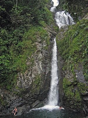 Waterfall in lush green mountains, man sitting on a rock on the bottom left, the waterfall is called Salto Doña Juana