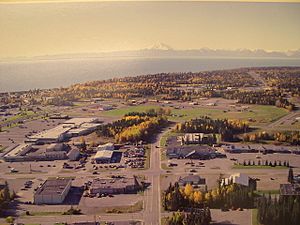 Aerial view of part of downtown Kenai.  The intersection of Willow Street and Barnacle Way is in the center of the foreground.  Cook Inlet and Mount Redoubt are in the background.