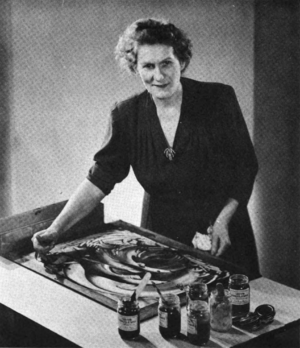 Ruth Faison Shaw standing by a finger painting on a table with paint jars, looking toward the camera
