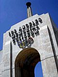 Photograph of the peristyle gate of the Los Angeles Memorial Coliseum, tile mosaicon the underside of the arch, and the Olympic torch reaching to the blue sky.