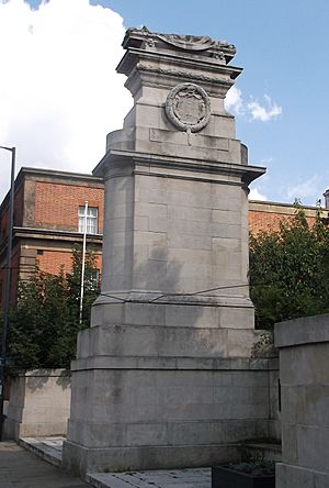 Side view of a war memorial in white stone. A coat of arms surrounded by a laurel wreath is carved under an effigy of a soldier.