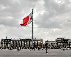 The Zócalo also known as Plaza de la Constitución with the Mexican flag waving in the center and to the right behind it, the Old Portal de Mercaderes